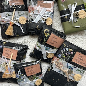 May include: Six rectangular gift packages wrapped in black and olive green paper with floral and celestial patterns. Each package features a brown paper label with handwritten text, a white ribbon, a small metal pentacle charm, and a crystal wand.
