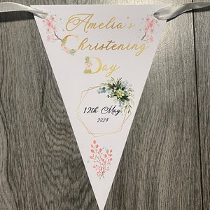 May include: A white triangular banner with gold lettering that reads "Amelia's Christening Day 12th May 2024". The banner is decorated with pink and green floral illustrations.