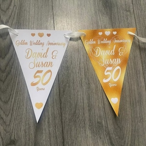 May include: Two white and gold triangular pennants with gold hearts and the text "Golden Wedding Anniversary David & Susan 50 Years" on each.