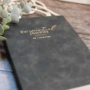May include: A dark gray planner with a matte finish featuring the gold embossed text "the essential planner" and "A5 - UNDATED" on the front cover. The rectangular book has a clean, solid-colored surface and is positioned at an angle.