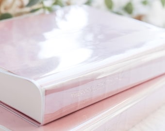 Puede incluir: Dos libros apilados de color rosa claro con lomos blancos y el texto "THE COZY HOME" en una fuente delicada. Los libros están envueltos en plástico transparente, reflejando la luz. El fondo está borroso con vegetación.