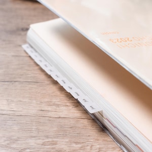 Könnte beinhalten: Ein Planer mit cremefarbenem Einband und dem orangefarbenen Text "HOBONICHI 2023 cousin". Der Planer verfügt über weiße Registerkarten an der Seite, die jeweils mit den Monaten des Jahres in schwarzer Schrift beschriftet sind, beginnend mit Jan, Feb, Mar, Apr, May.