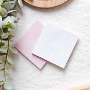 May include: Two square notepads, one pink and one white, with a grid pattern. The notepads are on a white, textured surface, with a sprig of greenery in the upper left corner. A wooden tray is visible in the background.