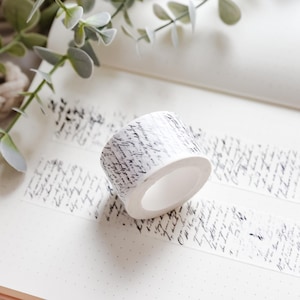 May include: A roll of decorative washi tape featuring a white base and black script. The tape is partially unrolled, showing a strip of the same design. The tape rests on a dotted paper surface, with greenery in the background.