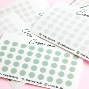 May include: A collection of white sticker sheets, each covered in rows of light green and grey dots, are displayed on a pink background. The sheets feature the text "Cosmicaa" and social media details. These are craft stickers.