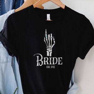 May include: Black t-shirt featuring a white skeleton hand wearing a ring and the text "Bride or Die".