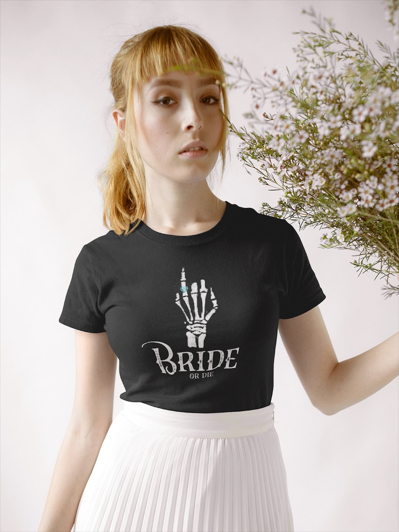May include: A black t-shirt featuring a white skeleton hand wearing a ring. The text "Bride or Die" is printed below the hand.