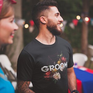 May include: A black t-shirt with a floral design and the word "groom" printed in white. The design features a spiderweb with flowers and leaves.
