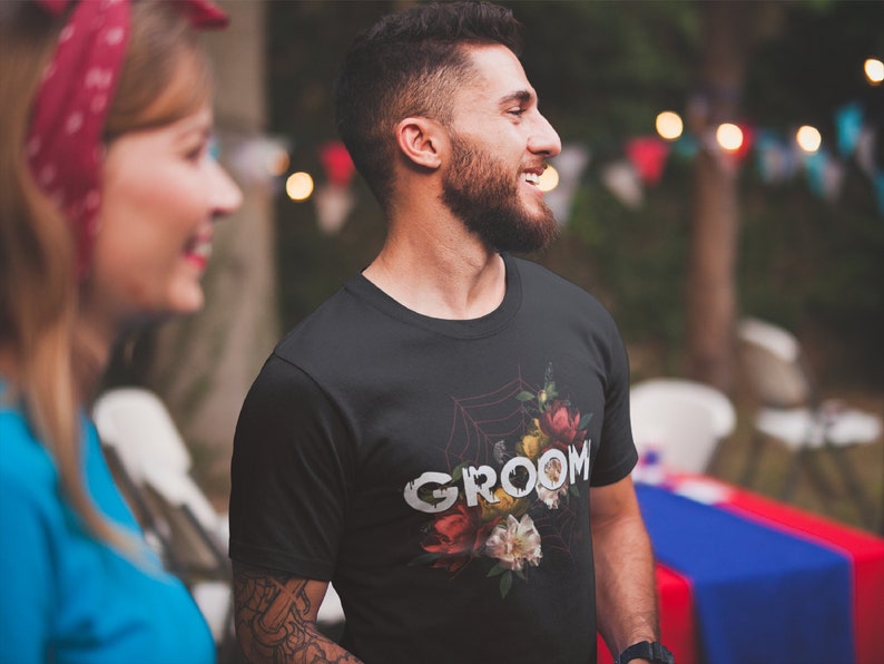 May include: A black t-shirt with a floral design and the word "groom" printed in white. The design features a spiderweb with flowers and leaves.