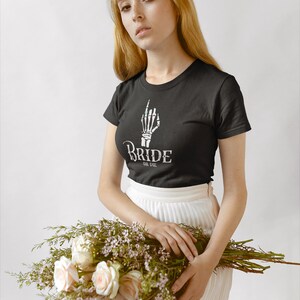 May include: A woman wearing a black t-shirt with a skeleton hand graphic and the text "Bride or Die" is holding a bouquet of white roses and greenery. She is wearing a white skirt.
