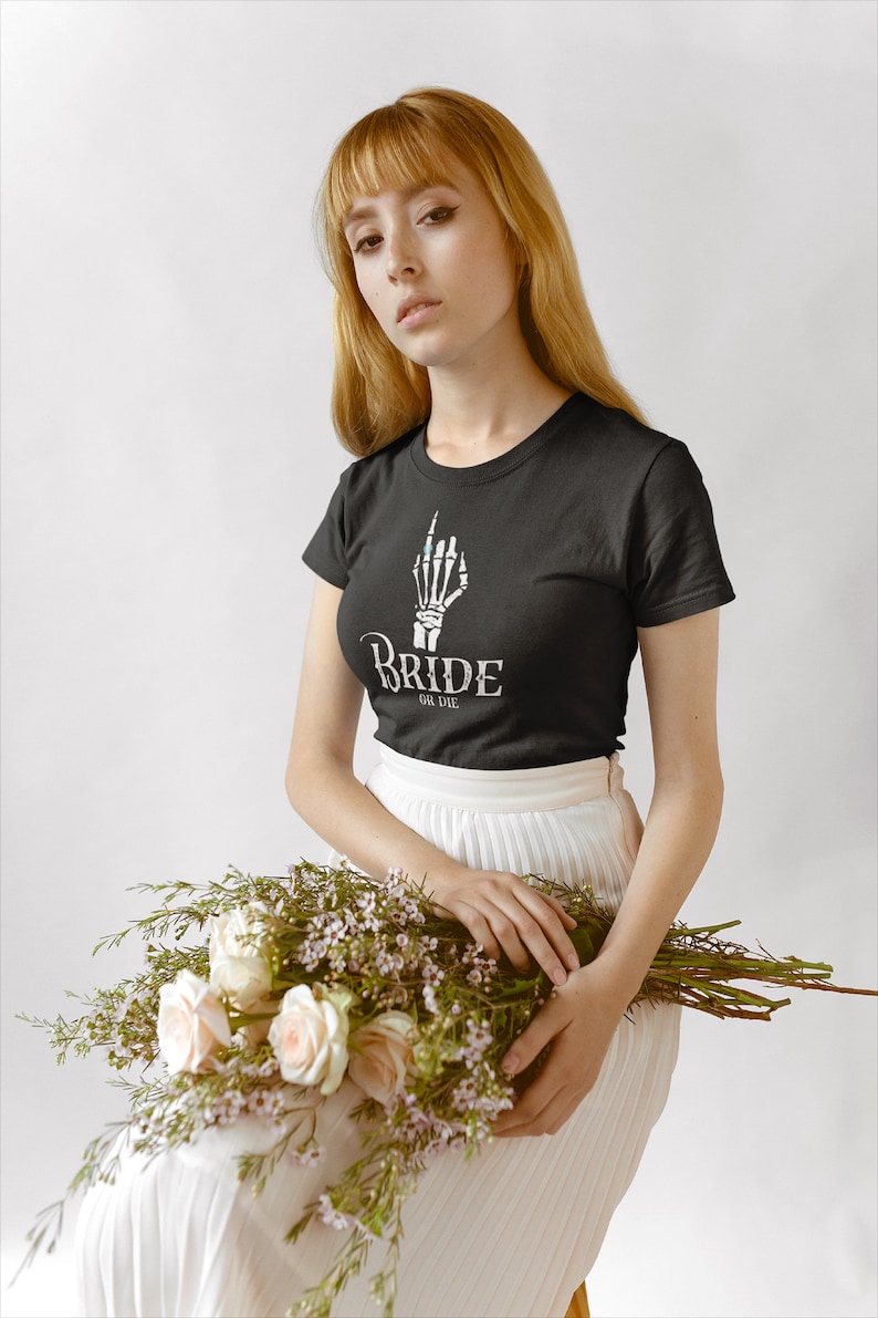 May include: A woman wearing a black t-shirt with a skeleton hand graphic and the text "Bride or Die" is holding a bouquet of white roses and greenery. She is wearing a white skirt.