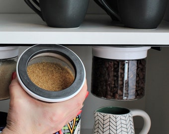 May include: A kitchen shelf with two black mugs, two glass canisters with lids, and a white ceramic mug with a colourful heart design. The canisters are filled with brown sugar and coffee beans. The shelf is made of white wood.