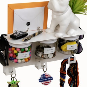 Può includere: Organizzatore da parete bianco con tre barattoli trasparenti etichettati "Treats", "Change" e "Keys". L'organizzatore ha ganci per appendere chiavi, occhiali da sole e una fascia per capelli. Una statuetta in ceramica bianca di un bulldog francese è posizionata sopra l'organizzatore. Una busta arancione è fissata alla parte superiore dell'organizzatore.