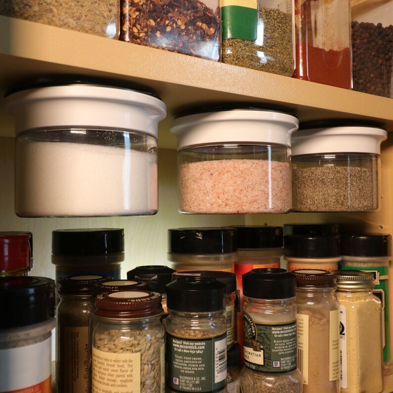 May include: A close-up of a kitchen cabinet with various spices and seasonings in glass jars. The jars are organized on shelves and some are hanging from the top of the cabinet. The jars are filled with different types of spices, including salt, pepper, and herbs.