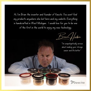May include: A man with a blue shirt is leaning forward, looking at the camera. He is sitting at a table with six glass jars with black lids. The jars are filled with various grains and spices. The text "vascito" is visible in the lower right corner of the image.