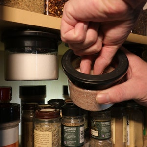 May include: A hand reaches into a spice rack to grab a jar of pink Himalayan salt. The jar is clear glass with a black lid and a black ring around the top. The salt is a light pink color. The spice rack is filled with various other jars of spices.