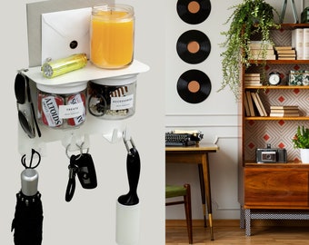Puede incluir: Organizador blanco de pared con ganchos para colgar llaves, un paraguas negro y un rodillo para pelusas. Hay dos frascos de vidrio con tapas, uno con la palabra "Treats" y el otro con la palabra "Accessories". Un sobre blanco está en el estante superior con una vela amarilla y una linterna plateada.