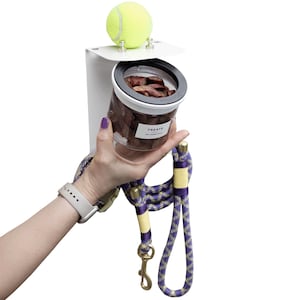 Puede incluir: Un organizador de pared blanco con un estante superior que sostiene una pelota de tenis amarilla, un frasco de pl&aacute;stico transparente lleno de premios marrones con la etiqueta TREATS y una correa trenzada morada y amarilla con un gancho dorado.