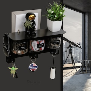 Op de afbeelding: Zwarte wandmontage sleutelhanger met drie haken, drie glazen potten met de labels "Change", "Altoids Treats" en "Accessories", en een klein astronautbeeldje. De sleutelhanger heeft ook een kleine groene Yoda sleutelhanger en een sleutelhanger met de tekst "American Made".