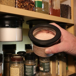 May include: A hand holds a clear glass jar with a black lid, filled with pink Himalayan salt. The jar is being removed from a shelf with other spice jars.