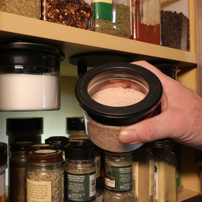 May include: A hand holds a clear glass jar with a black lid, filled with pink Himalayan salt. The jar is being removed from a shelf with other spice jars.