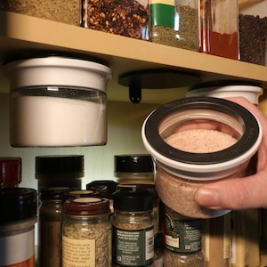 May include: A close-up of a variety of spices and seasonings in glass jars, some of which are hanging from the ceiling. The jars are filled with different types of salt, sugar, and other spices. The jars are all clear glass and have black lids.