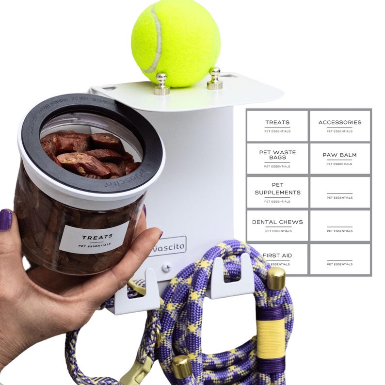 Puede incluir: Un organizador de pared blanco para mascotas con una pelota de tenis en la parte superior, un frasco transparente con la etiqueta TREATS y ganchos que sostienen una correa de cuerda con patr&oacute;n morado y amarillo. Una hoja de diez etiquetas.