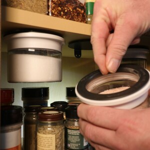 May include: A close-up of a hand reaching into a white and black spice jar with a clear glass container. The jar is attached to a magnetic base and is hanging from a wooden shelf. Other spice jars are visible on the shelf.