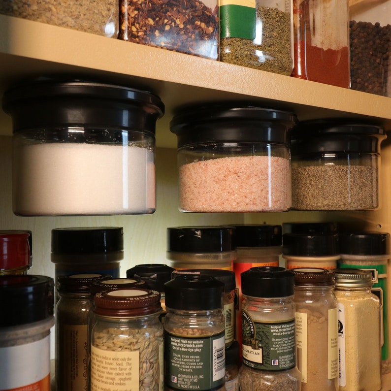 May include: A close-up of a spice rack with various spices in glass jars. The jars are organized on a wooden shelf and some are hanging from the shelf. The spices include salt, pepper, and other herbs and seasonings.