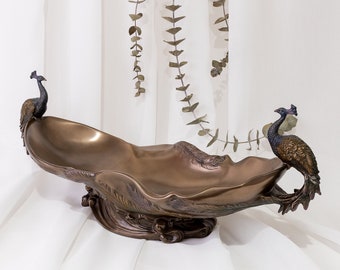 Puede incluir: Un cuenco decorativo de bronce con dos figuras de pavo real a cada lado. El cuenco tiene una forma ondulada, orgánica y un diseño detallado.