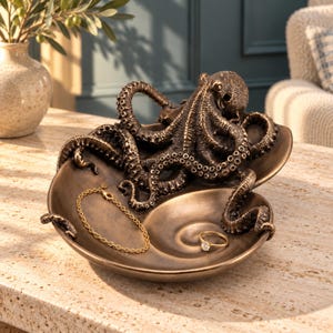 May include: A bronze-toned decorative bowl shaped like a seashell, featuring a detailed octopus sculpture resting on top. The bowl contains a delicate gold-colored chain bracelet and a simple gold ring with a small clear stone.