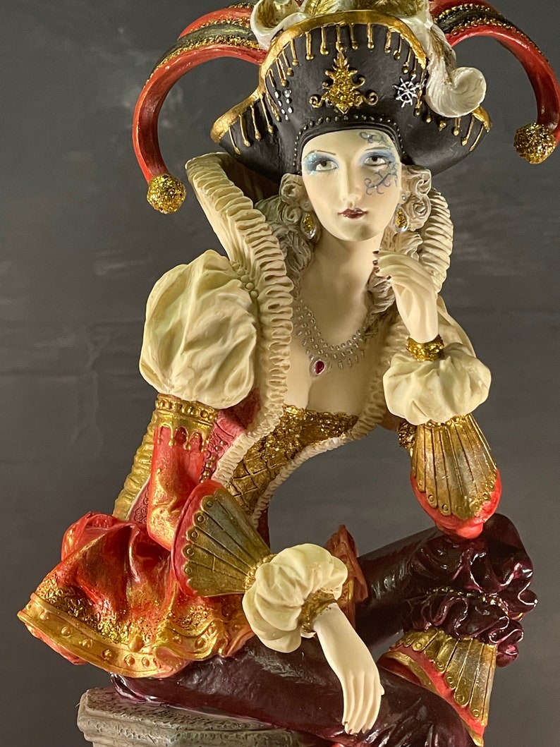 Puede incluir: Figura decorativa de un buf&oacute;n sentado. La figura lleva un sombrero negro con detalles dorados y ribetes rojos, un cuello con volantes y un atuendo con estampado rojo y dorado. La figura tiene una tez p&aacute;lida y est&aacute; adornada con joyas.