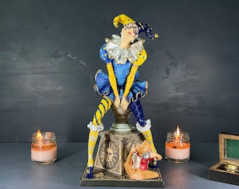Op de afbeelding: Gedetailleerd beeldje van een nar in een blauw, geel en wit kostuum. De nar poseert bovenop een decoratieve basis met een zonontwerp. Een klein teddybeerbeeldje zit aan de basis. Twee brandende kaarsen in glazen potten staan aan weerszijden.