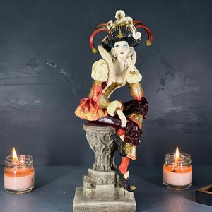 May include: A decorative figurine of a jester in a colourful costume, seated on a pedestal. The jester wears a black hat with red and gold accents. The figurine has a white ruffled collar and a red and gold patterned outfit.