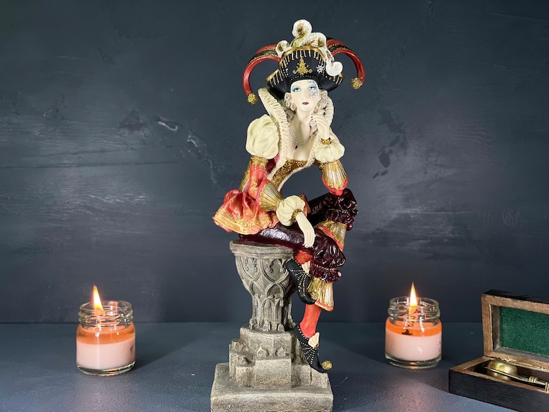 Puede incluir: Figura decorativa de un buf&oacute;n con un disfraz colorido, sentado en un pedestal. El buf&oacute;n lleva un sombrero negro con detalles rojos y dorados. La figura tiene un cuello blanco con volantes y un atuendo con estampado rojo y dorado.