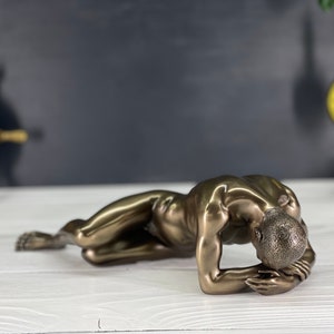 May include: A bronze sculpture of a person lying on their stomach with their head resting on their hands. The sculpture is made of a smooth, polished bronze and has a realistic appearance.
