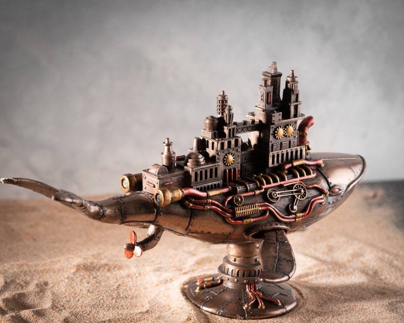 Op de afbeelding: Een steampunk-walvisbeeld met ingewikkelde details, met een miniatuurstad op zijn rug. De walvis is gemaakt van metaal en heeft een bronzen afwerking. Hij staat op een zanderig oppervlak.