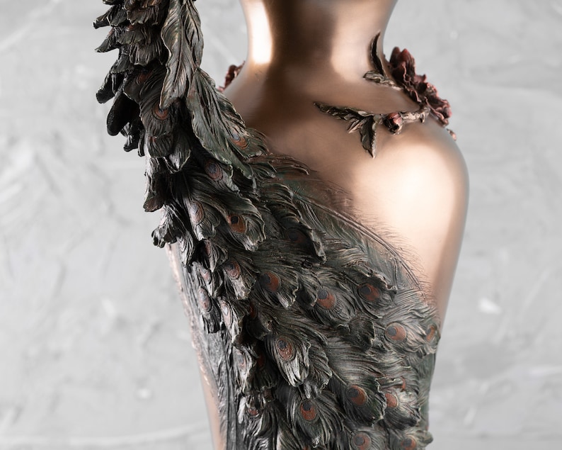 Puede incluir: Un jarr&oacute;n de color bronce con un dise&ntilde;o de pavo real. El jarr&oacute;n est&aacute; decorado con detalles intrincados, incluyendo plumas y un motivo floral.