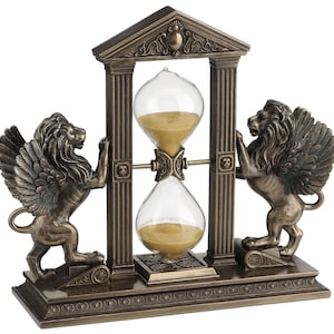 May include: A gold-toned hourglass with winged lion sculptures on each side, set on a decorative base with intricate details. The hourglass is framed by two pillars with ornate tops.