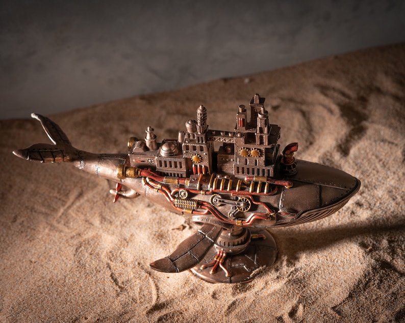 Op de afbeelding: Een steampunk walvisfiguur met ingewikkelde details, waaronder tandwielen, pijpen en een stadsachtige structuur op zijn rug. De walvis is gemaakt van metaal en heeft een bronzen afwerking. Hij staat op een zanderige ondergrond.
