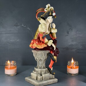 Puede incluir: Figura decorativa de una figura sentada con atuendo renacentista, con una combinaci&oacute;n de colores rojo, dorado y blanco. La figura est&aacute; posada sobre un pedestal detallado. La imagen tambi&eacute;n incluye dos velas encendidas en frascos de vidrio.