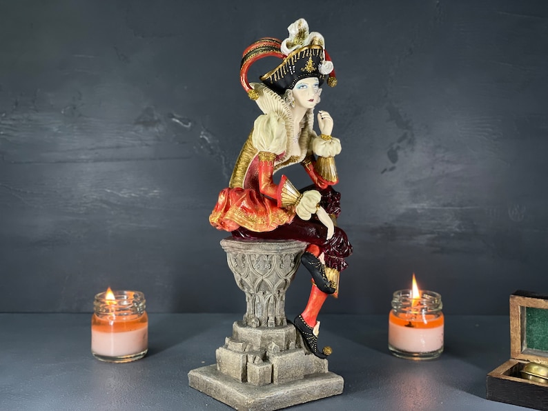 Puede incluir: Figura decorativa de una figura sentada con atuendo renacentista, con una combinaci&oacute;n de colores rojo, dorado y blanco. La figura est&aacute; posada sobre un pedestal detallado. La imagen tambi&eacute;n incluye dos velas encendidas en frascos de vidrio.