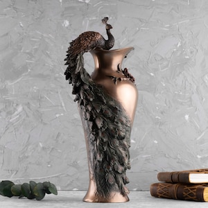 Puede incluir: Un jarr&oacute;n de color bronce con un dise&ntilde;o de pavo real. Las plumas de la cola del pavo real est&aacute;n extendidas y la cabeza del p&aacute;jaro est&aacute; posada en la parte superior del jarr&oacute;n.