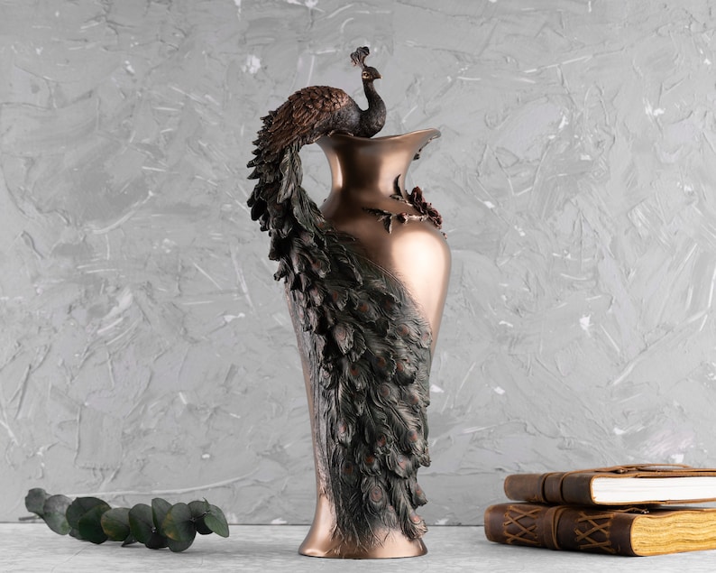 Puede incluir: Un jarr&oacute;n de color bronce con un dise&ntilde;o de pavo real. Las plumas de la cola del pavo real est&aacute;n extendidas y la cabeza del p&aacute;jaro est&aacute; posada en la parte superior del jarr&oacute;n.