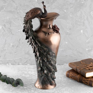Puede incluir: Un jarr&oacute;n decorativo de bronce con un dise&ntilde;o de pavo real. Las plumas de la cola del pavo real est&aacute;n intrincadamente detalladas y el jarr&oacute;n tiene un dise&ntilde;o de rosa en el lateral.
