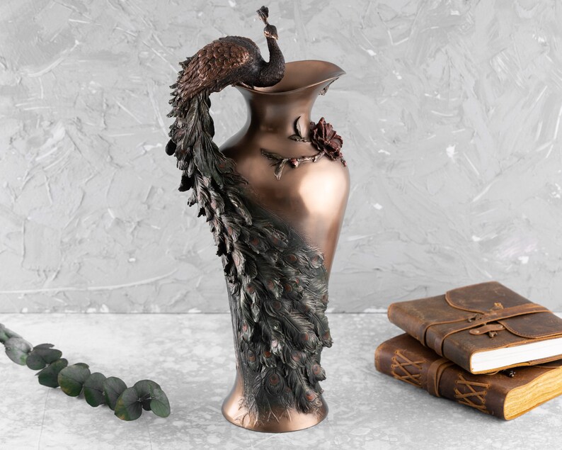 Puede incluir: Un jarr&oacute;n decorativo de bronce con un dise&ntilde;o de pavo real. Las plumas de la cola del pavo real est&aacute;n intrincadamente detalladas y el jarr&oacute;n tiene un dise&ntilde;o de rosa en el lateral.