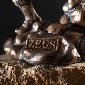 K&ouml;nnte beinhalten: Bronzeskulptur von Zeus, dem griechischen Gott des Himmels und des Donners, stehend auf einem felsigen Sockel. Die Skulptur ist detailliert und tr&auml;gt eine Inschrift auf dem Sockel, die "ZEUS" lautet.