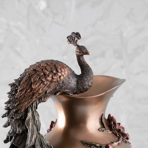 Puede incluir: Un jarr&oacute;n decorativo de bronce con un dise&ntilde;o de pavo real. El pavo real est&aacute; posado en la parte superior del jarr&oacute;n y tiene la cabeza girada hacia un lado. El jarr&oacute;n est&aacute; decorado con detalles florales.