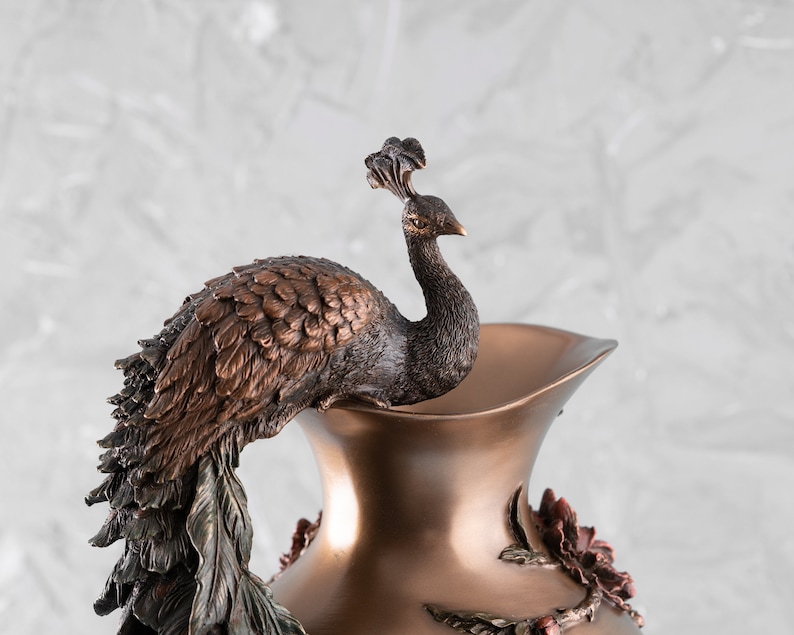 Puede incluir: Un jarr&oacute;n decorativo de bronce con un dise&ntilde;o de pavo real. El pavo real est&aacute; posado en la parte superior del jarr&oacute;n y tiene la cabeza girada hacia un lado. El jarr&oacute;n est&aacute; decorado con detalles florales.
