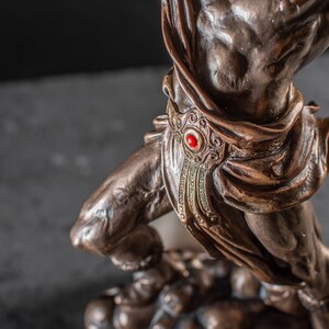 K&ouml;nnte beinhalten: Bronzeskulptur einer Kriegerfigur mit einem roten Juwel am G&uuml;rtel. Der Krieger steht auf einem Sockel mit anderen Figuren.